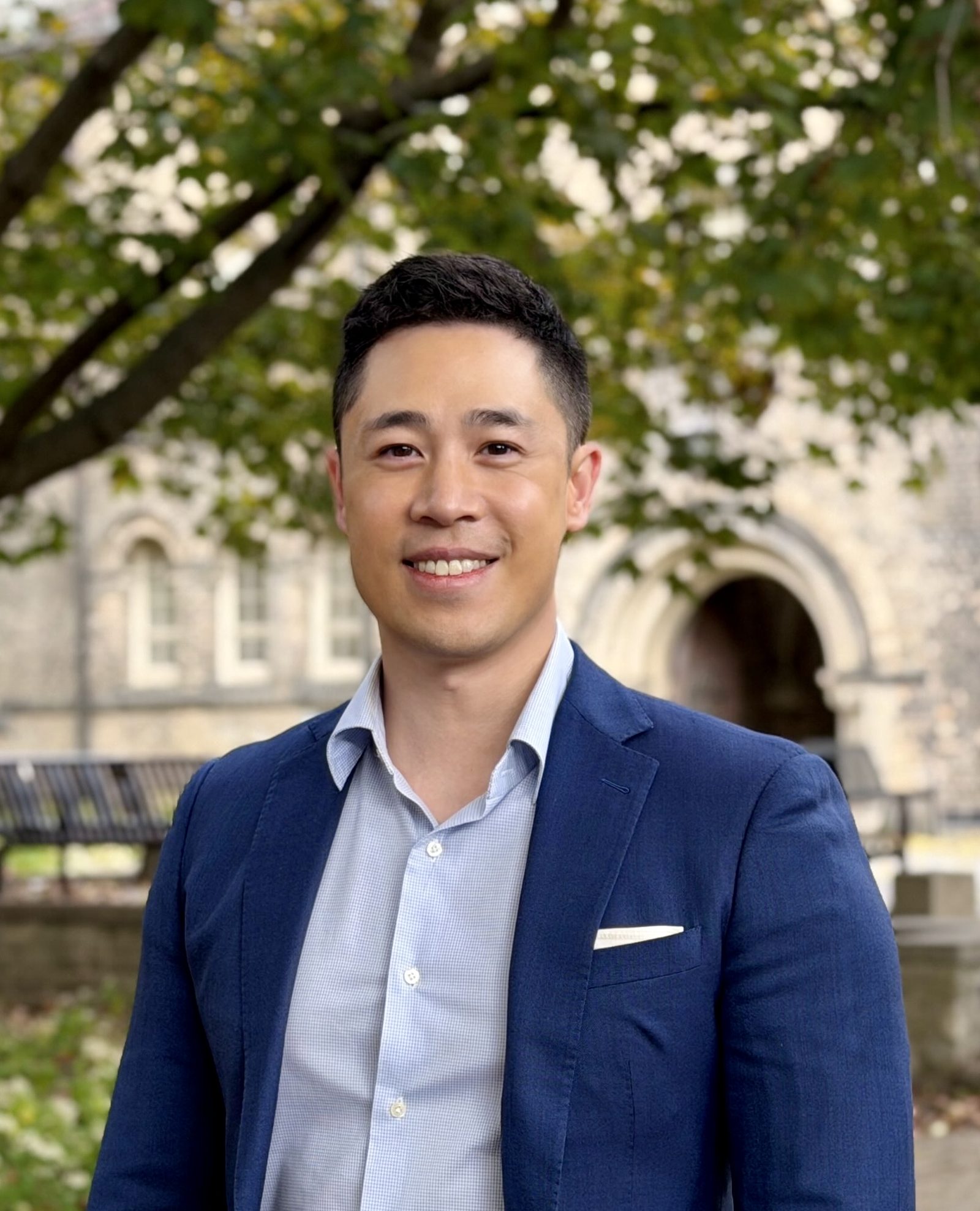 Victor Wong standing beneath green maple foliage in front of a historic stone building at the University of Toronto.