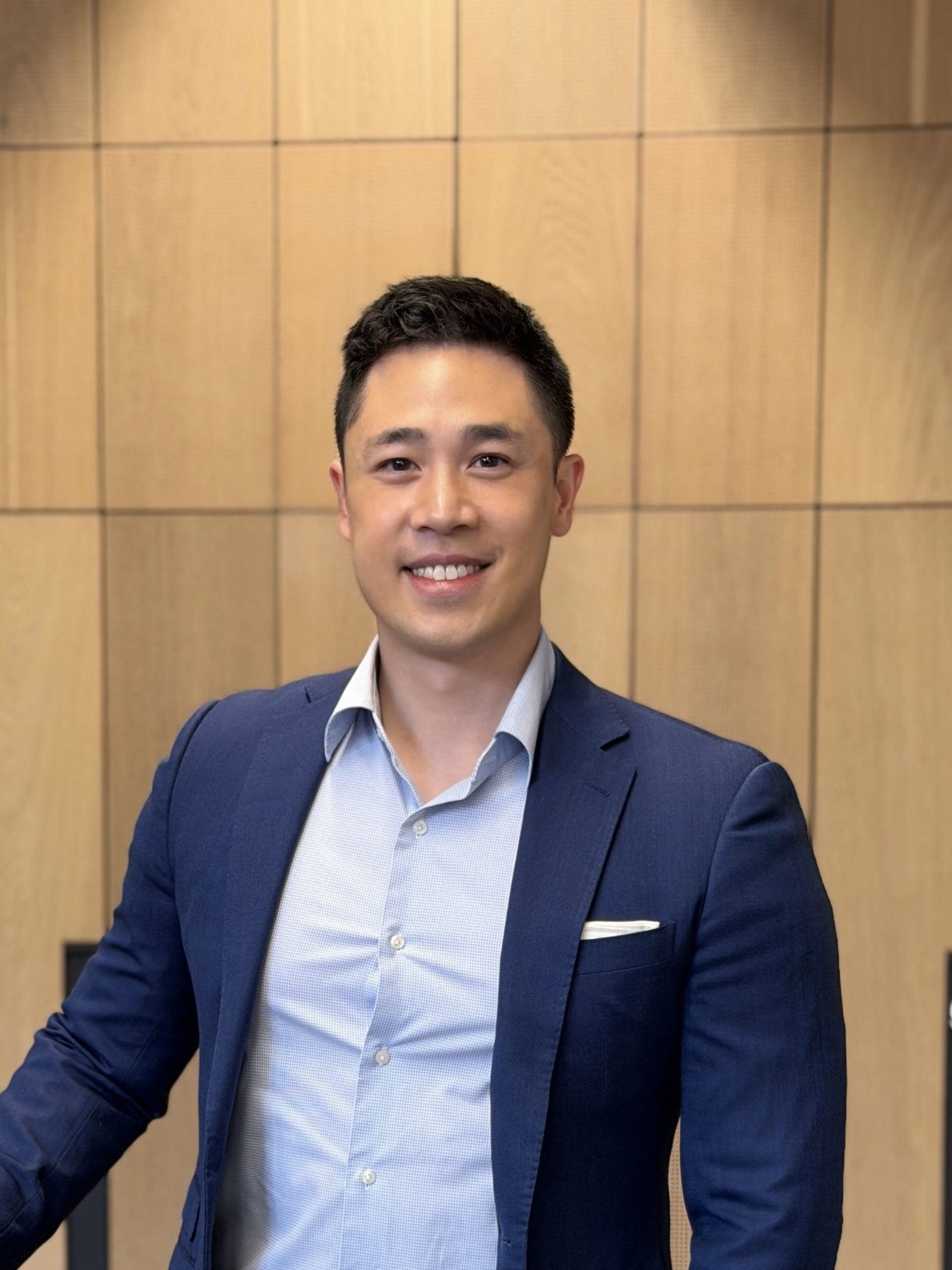Victor Wong, pharmacist, smiling warmly in a navy blazer and light-blue shirt against oak wood panelling.